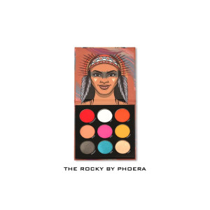 Open makeup palette with nine circular eyeshadows in red, white, orange, peach, pink, yellow, charcoal, teal, and beige; lid features a stylized portrait of a Native American person wearing a feathered headband.