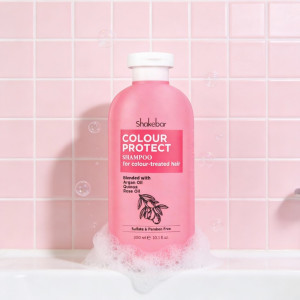 Pink shampoo bottle labeled Color Protect for colour-treated hair on a bubbly sink in a pink-tiled bathroom.