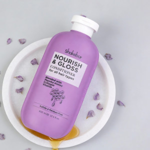 Purple Shakebar Nourish & Gloss Conditioner bottle on a white plate with scattered petals and honey spill, promoting hair nourishment for all hair types.